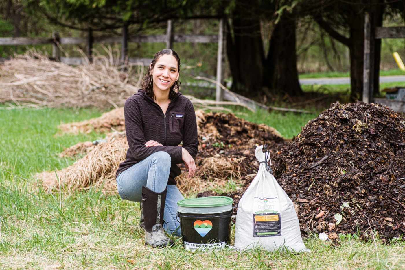 Hope LaBonty kneeling at the compost piles at Loving Earth Compost with a branded bucket and bag of finished compost