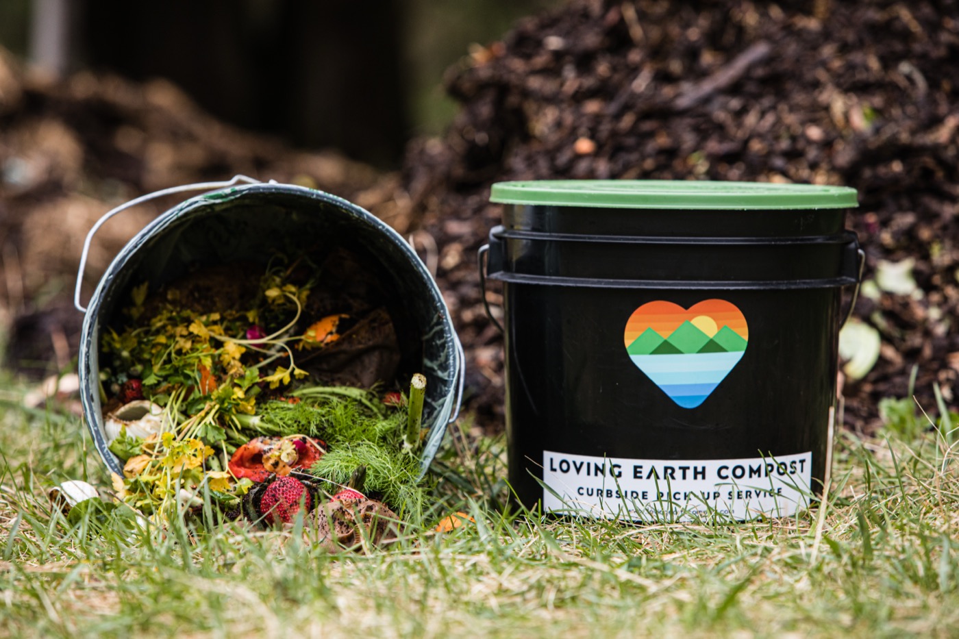 A Loving Earth Compost curbside bucket with the heart logo beside a tipped bucket of food scraps, compost pile behind